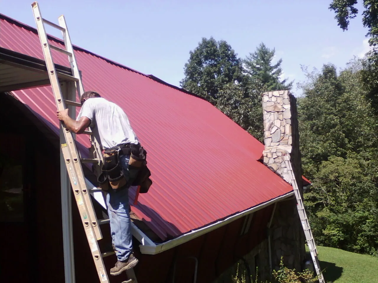 Roofer on a ladder beside a red metal roof and stone chimney — Soffit & Fascia Repair by Roofing Brothers in Micaville